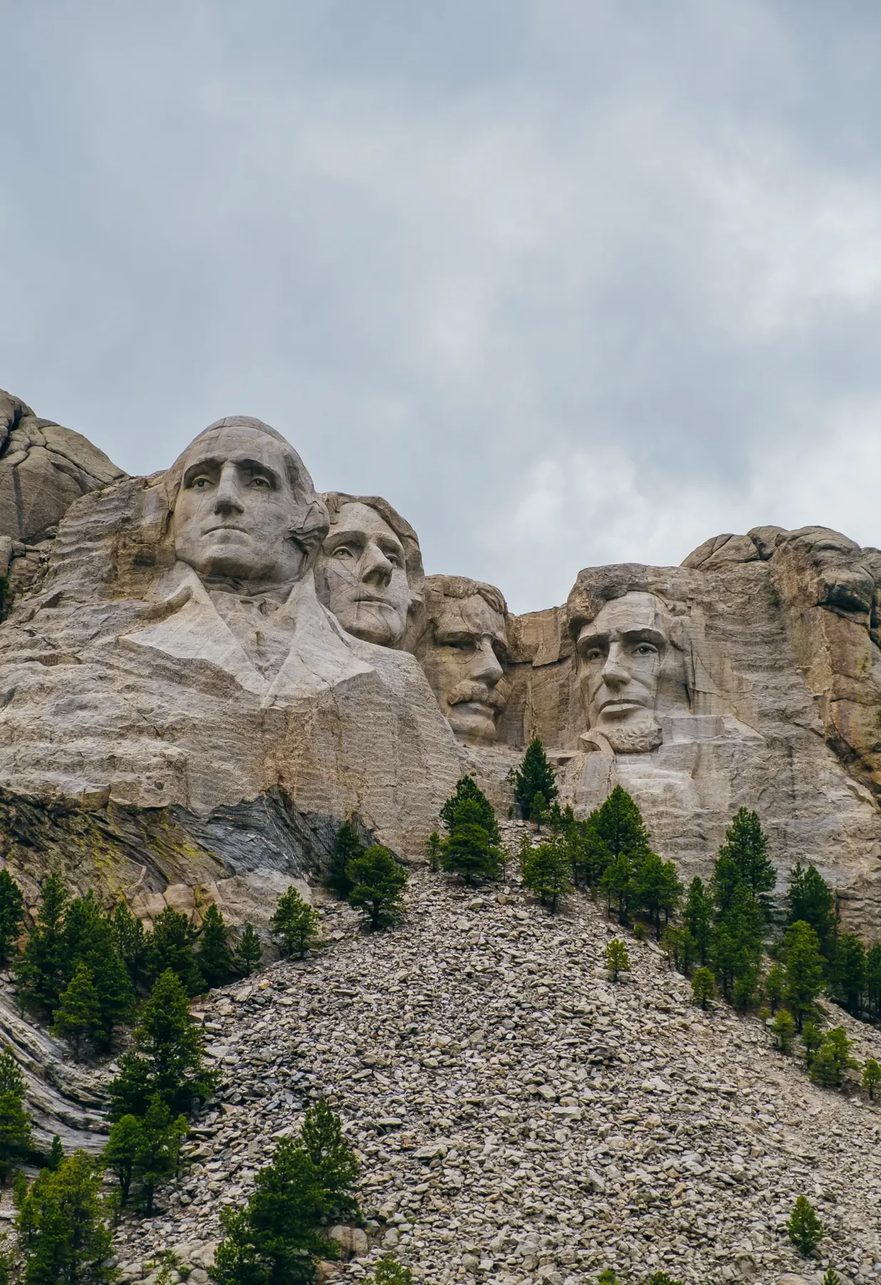 Mount Rushmore memorial above a slope of pines under an overcast sky.