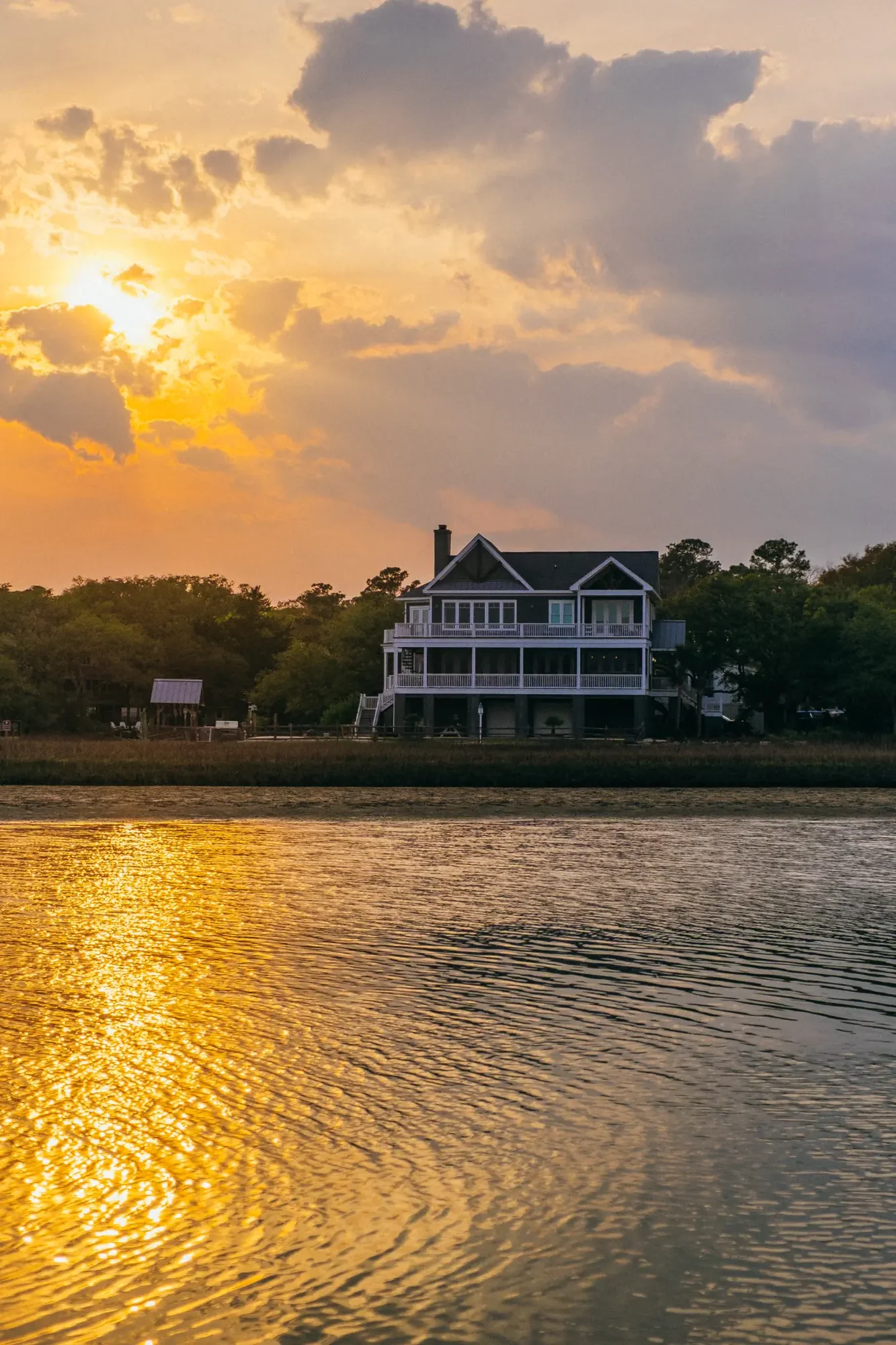 Large waterfront house silhouetted under warm golden sunset sky and glowing water reflection.