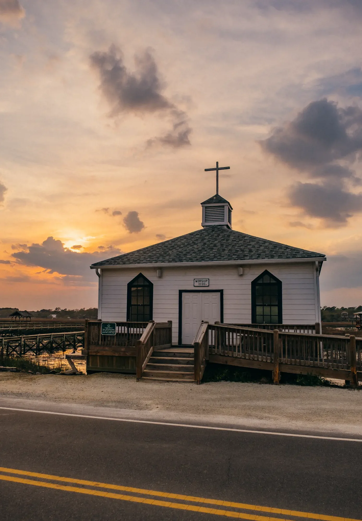 Small white building topped by a cross at golden sunset over marshland.