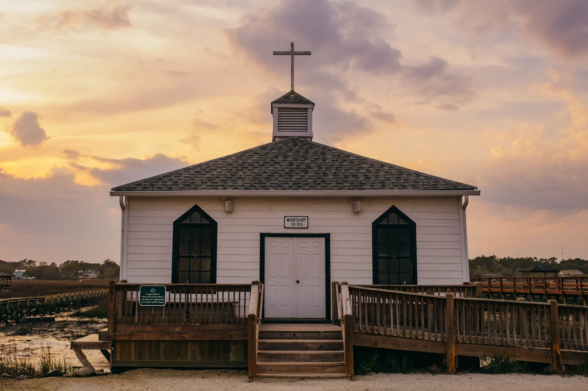 Small white chapel with cross on cupola above wooden ramp at golden sunset.