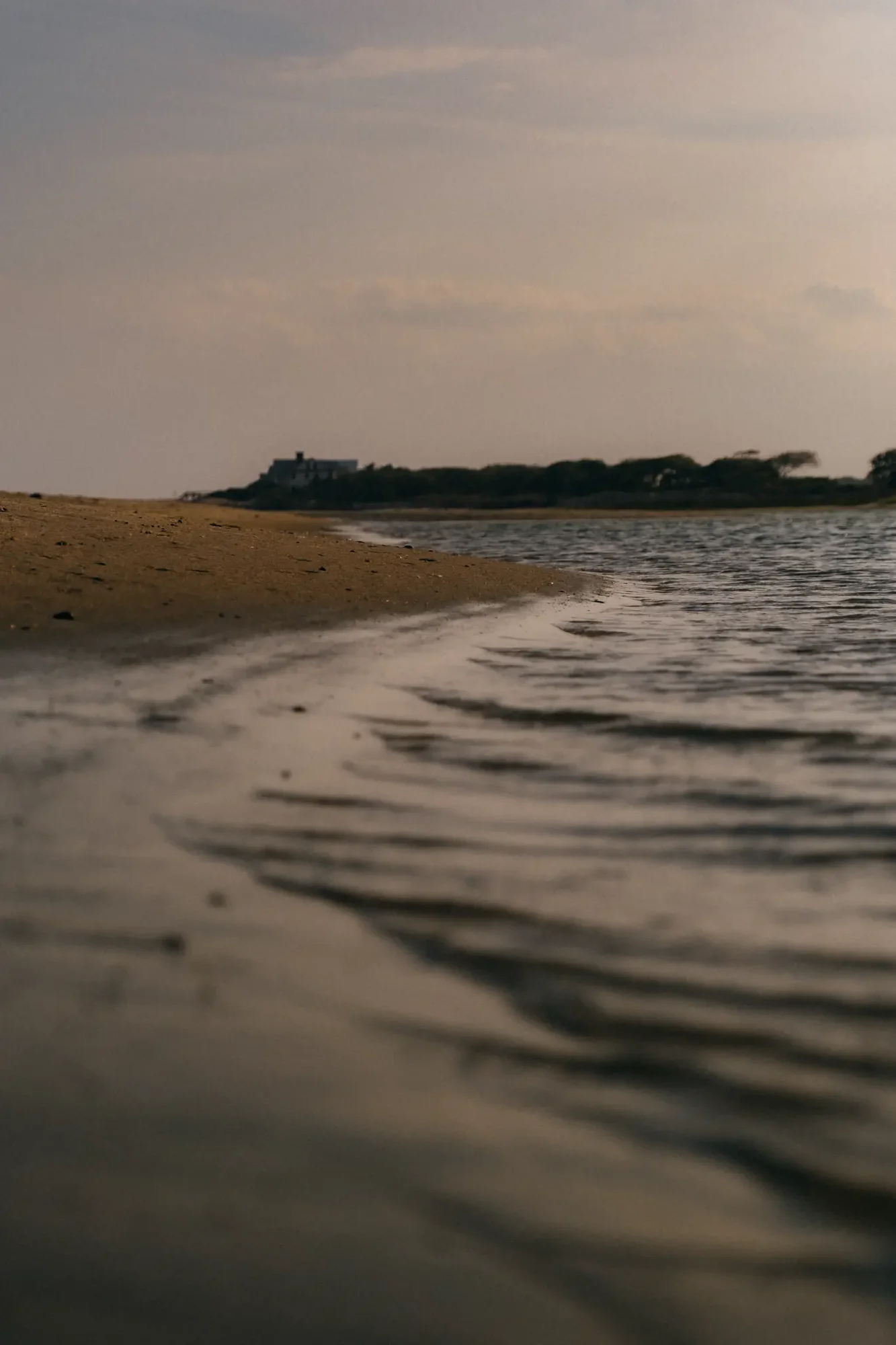 Low angle shoreline with rippling water foreground sandy beach leading to distant house silhouette at dusk.