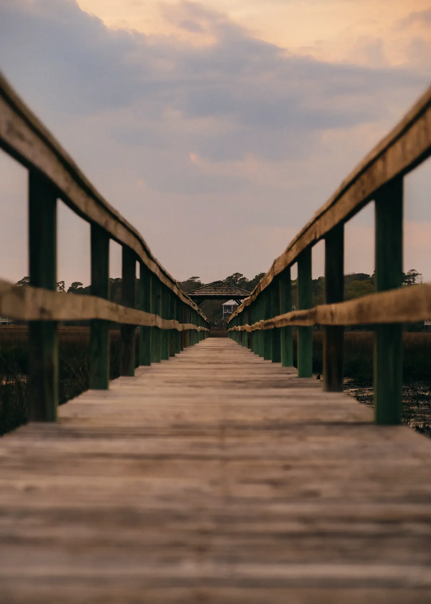Low wooden boardwalk with railings leading to distant gazebo under pastel sunset sky.