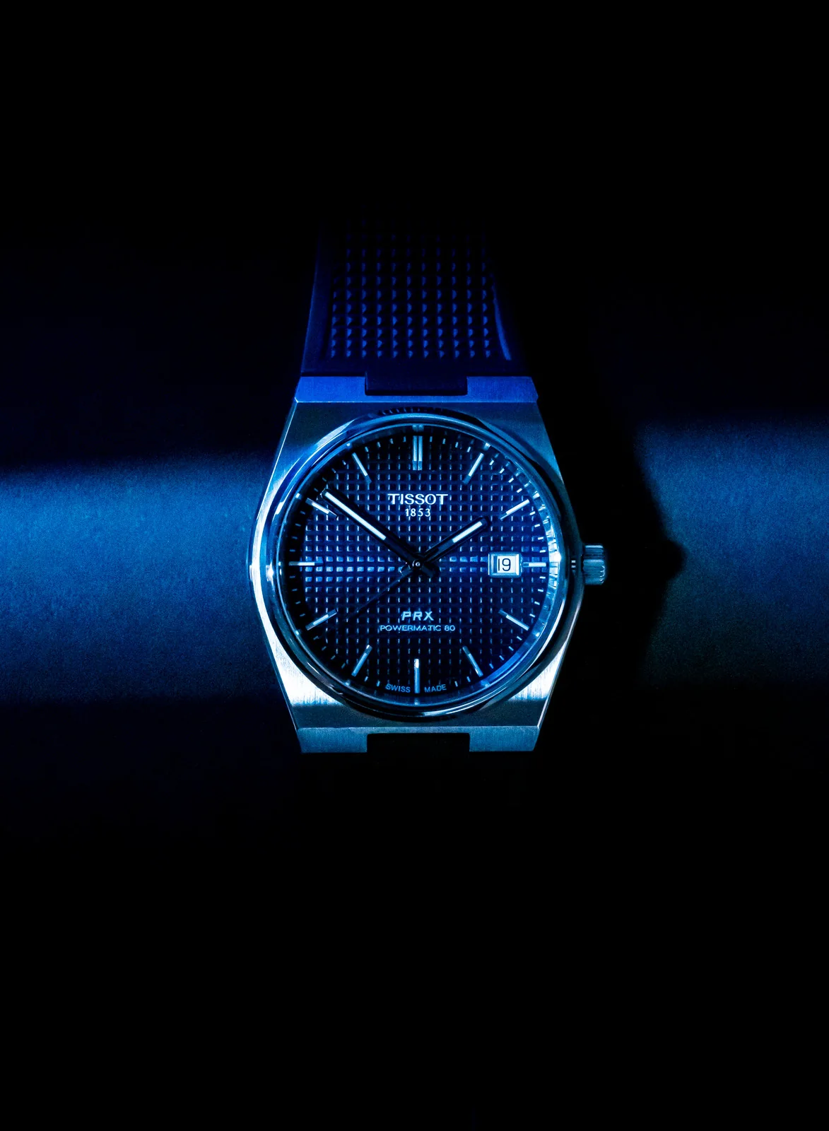 Blue-lit Tissot PRX watch with a dark textured dial and black rubber strap.