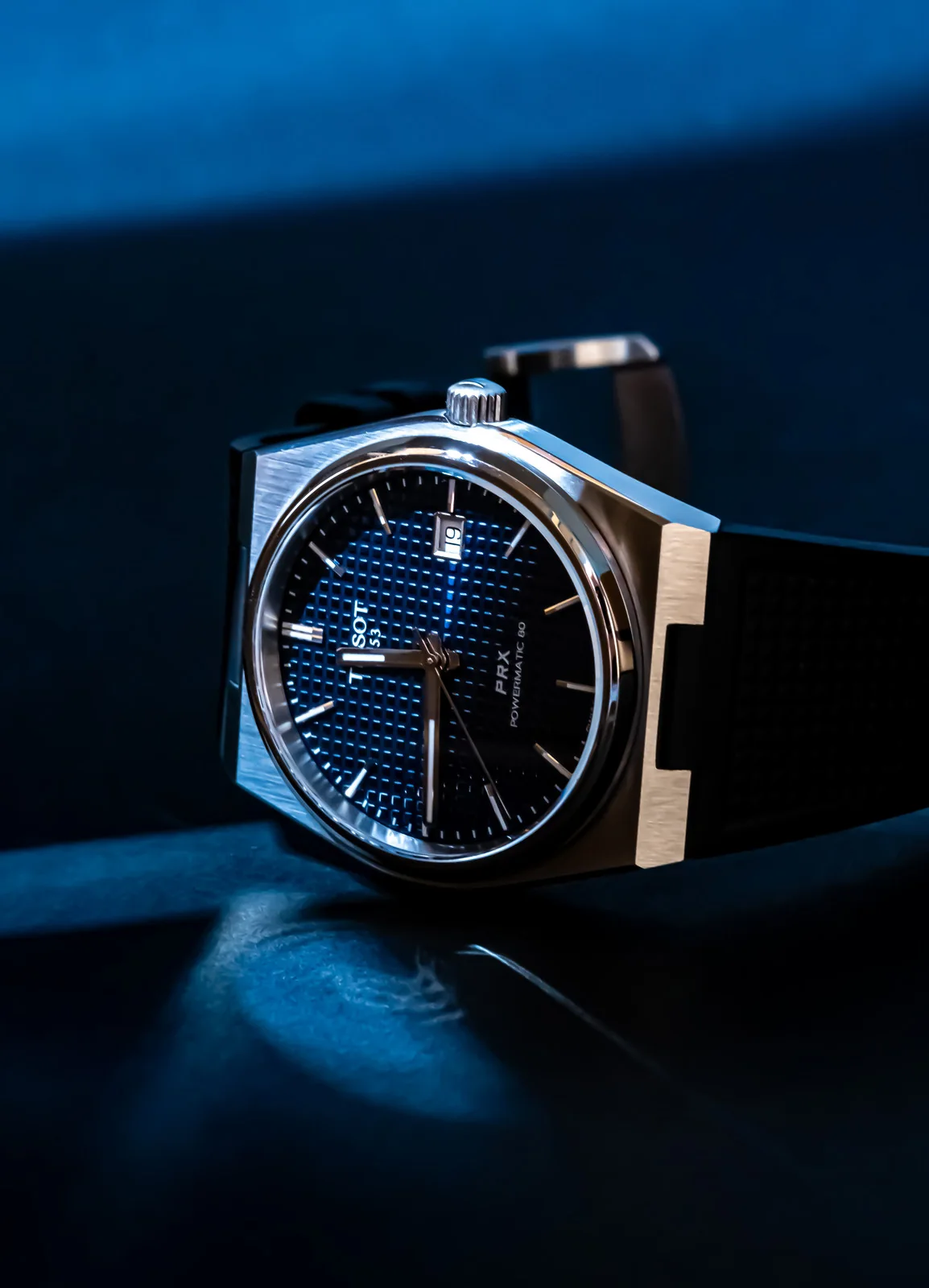 Tissot PRX watch photographed on dark fabric with a metallic blue-black dial.