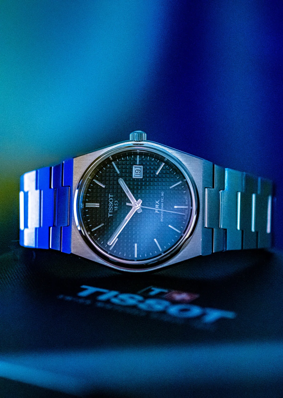 Tissot PRX watch illuminated by blue light above a Tissot wordmark.