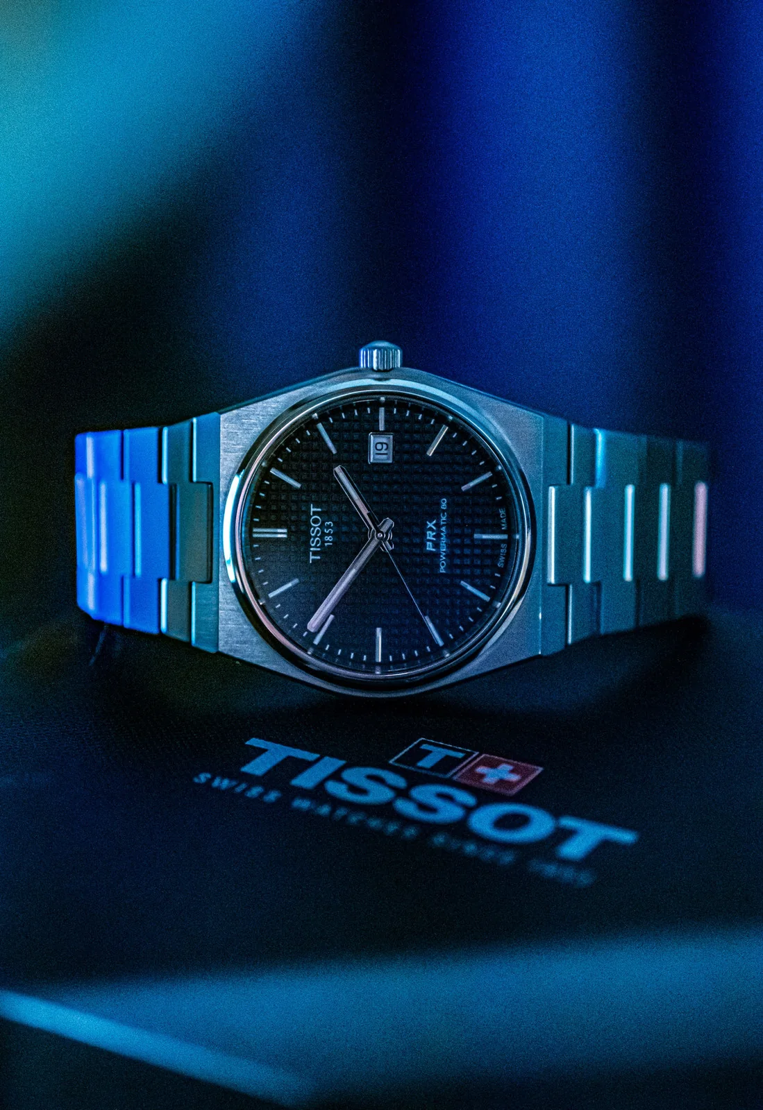 Tissot PRX watch on a blue surface with the Tissot wordmark below.